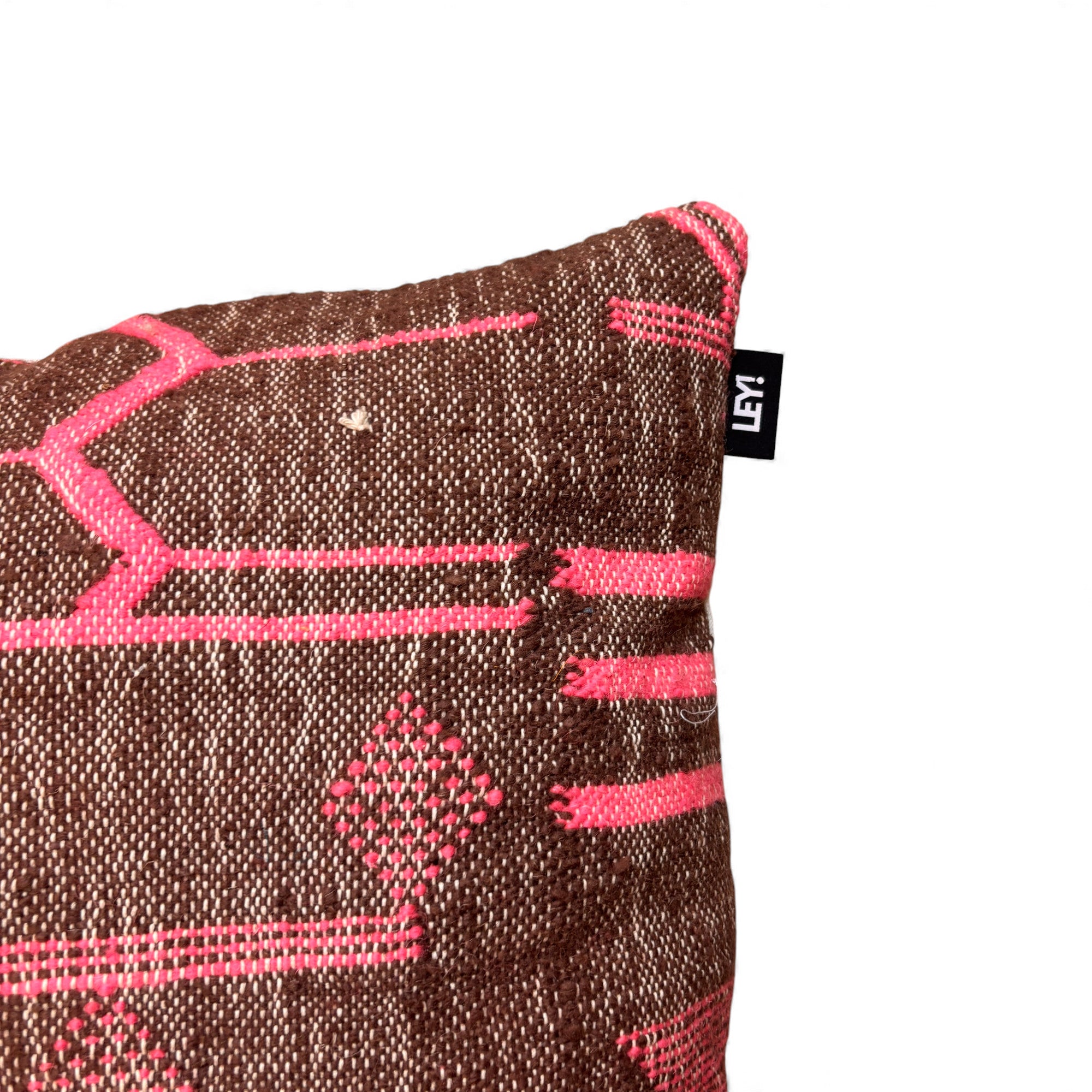 Eine Nahaufnahme des LEY! Kissen - Colorful Line - Fushia Edition 60x60 von LEY! aus brauner Wolle mit leuchtend rosa geometrischen Mustern und einem kleinen schwarzen Anhänger mit weißem Text an der Seite.