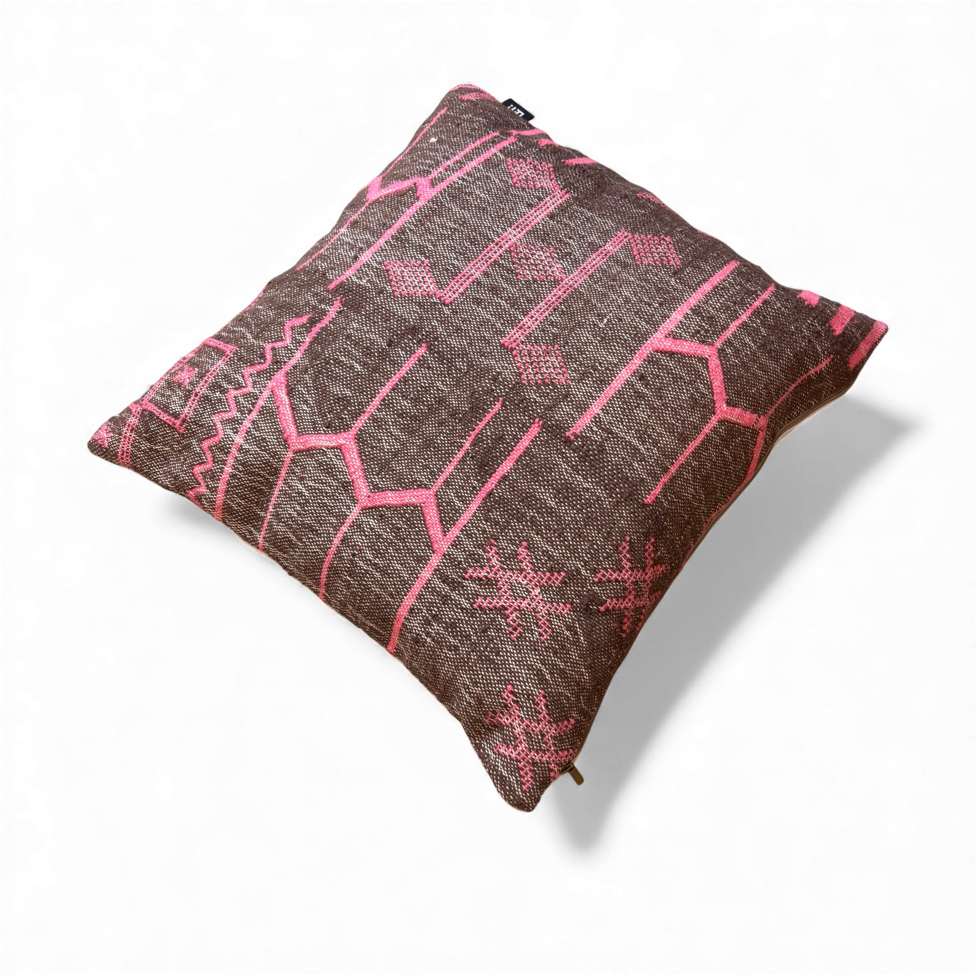 Das LEY! Kissen - Colorful Line - Fushia Edition 60x60 von LEY! ist ein quadratisches braunes Kissen mit rosa geometrischen und Tribal-Mustern, handgefertigt in Marrakesch, mit strukturiertem Stoff und einem kleinen schwarzen Eckanhänger, abgebildet auf weißem Hintergrund.
