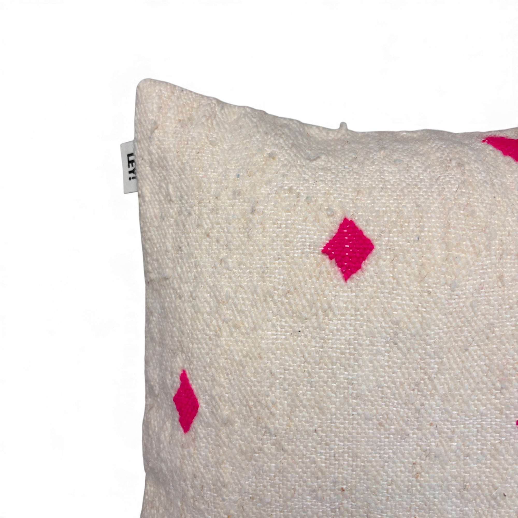 Eine Nahaufnahme des LEY! Kissen - Mona Pink - 50 x 50: ein strukturiertes, cremefarbenes Wollkissen mit hellrosa Rautenmustern und einem kleinen Anhänger an der Seite, handgefertigt nach Handarbeit Marrakesch, auf weißem Hintergrund.