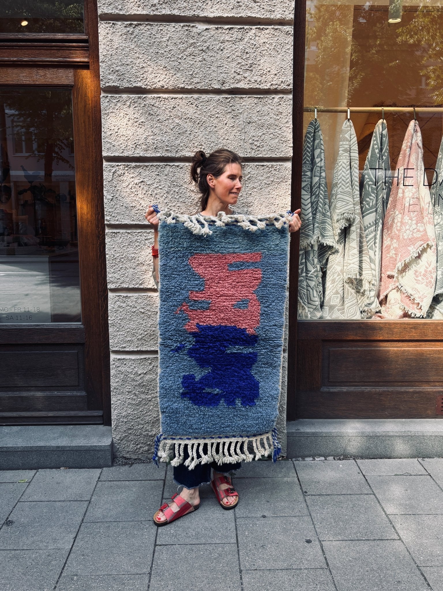 Eine Frau auf dem Bürgersteig hält vor einem Schaufenster einen „THE DANISH GIRL INTERIOR Berber-Teppich – Amani“ in den Händen.