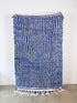 Der Berber Teppich - Blu - 1,20 x 2,00 von LEY! aus Schurwolle mit weißen und dunkelblauen Streifen, rauer Oberfläche und weißen Fransen hängt an einer schlichten weißen Wand.