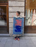 Eine Frau präsentiert den Berber-Teppich „THE DANISH GIRL INTERIOR“ – Amani, 0,60 x 1,10 m, mit blauen und rosa Mustern auf dem Bürgersteig.