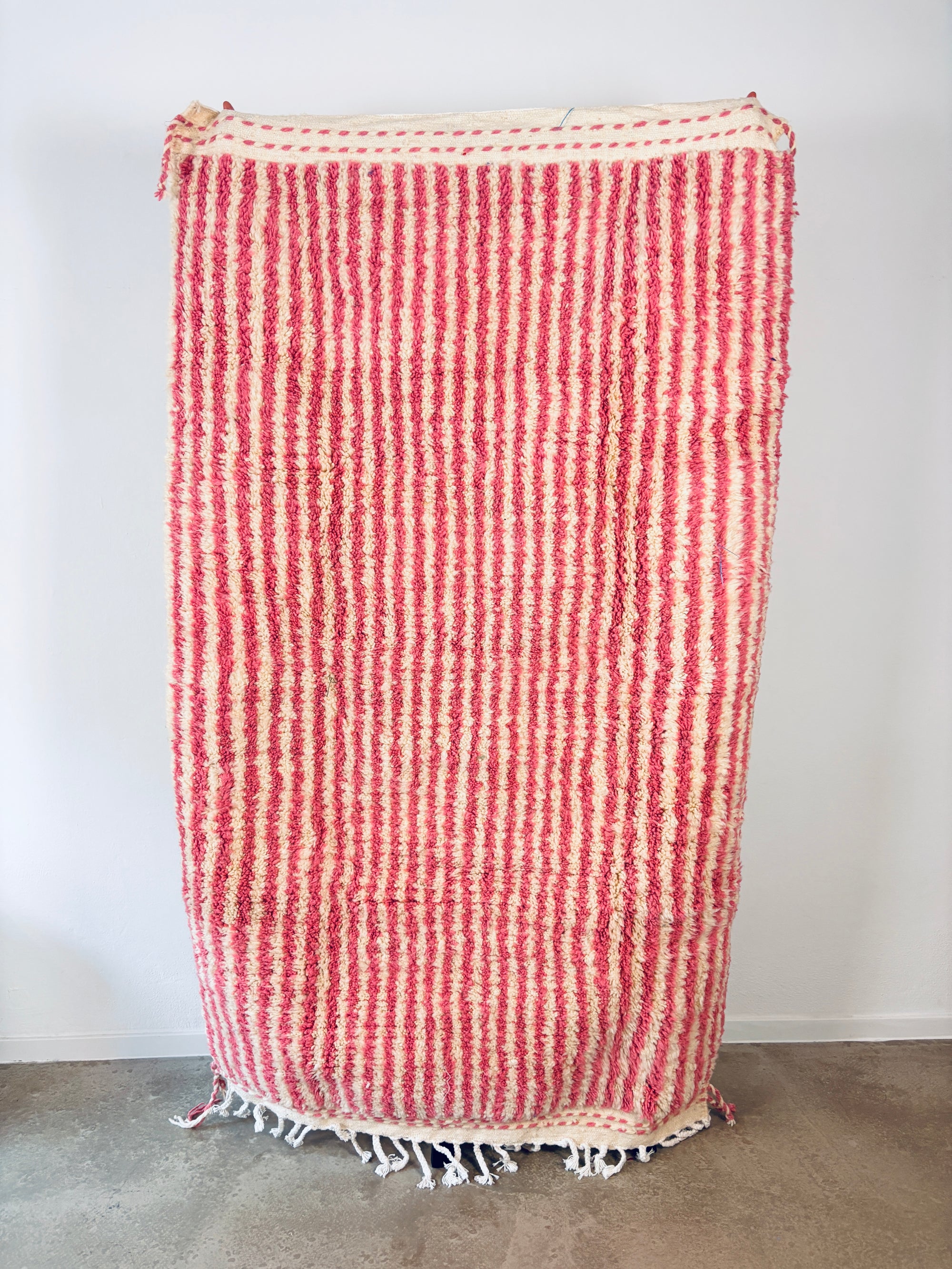 Der LEY! Berber Teppich - Pinq (1,20 x 2,00), handgewebt aus Schurwolle mit rosa-weißen Streifen und Fransen, hängt an einer weißen Wand - seine flauschige Struktur verleiht Charme und Wärme.