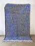 Der LEY! Berber Teppich - Blu (1,50 x 2,50) hängt senkrecht an einer weißen Wand, handgefertigt mit blauen und weißen Längsstreifen und mit weißen Fransen am unteren Ende auf einem hellgrauen Betonboden.
