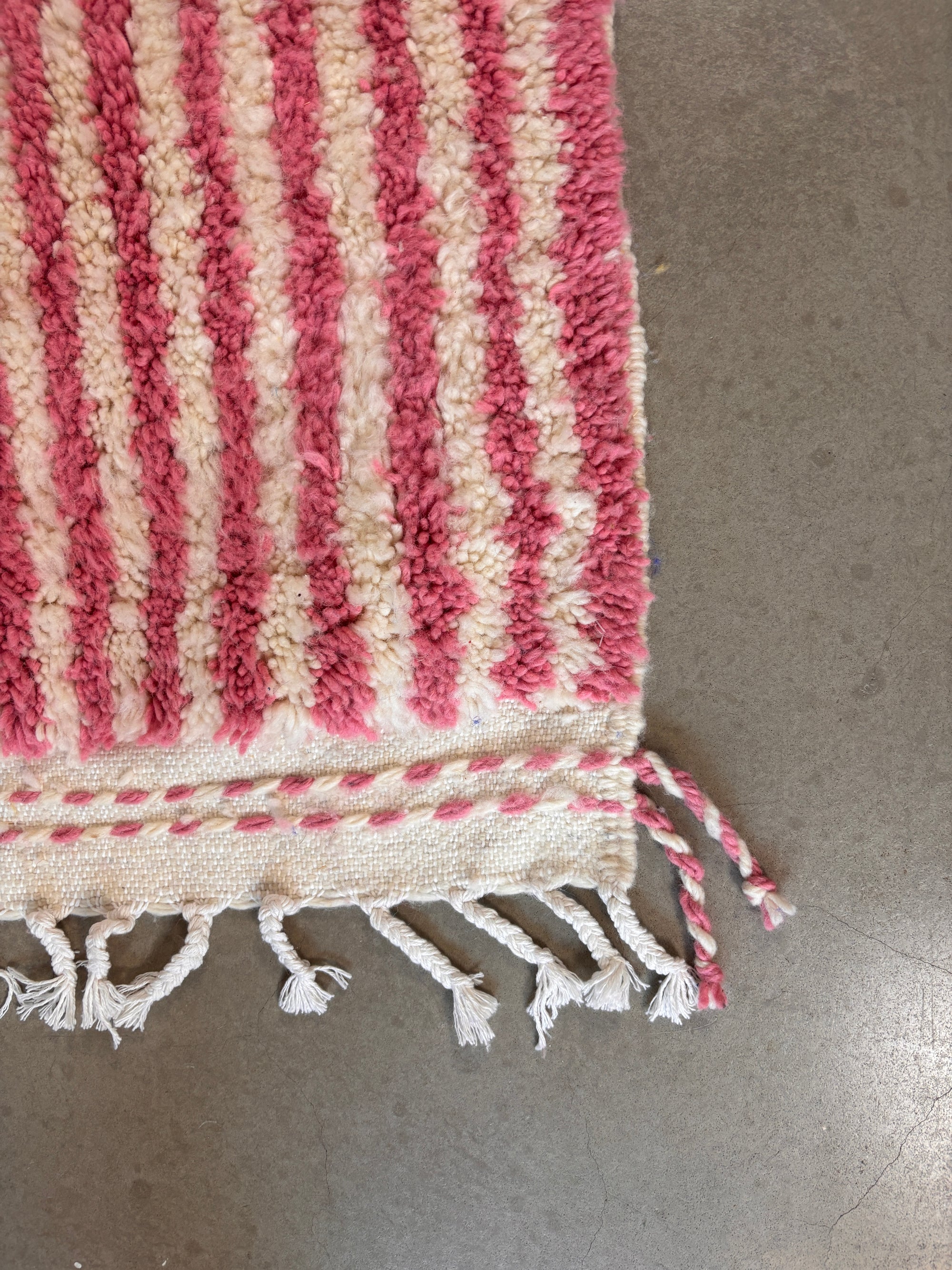 Eine Nahaufnahme des LEY! Berberteppich - Pinq (1,20 x 2,00), rosa und weiß gestreift mit geflochtenen Quasten an einem Rand, ausgestellt auf einem glatten grauen Boden.
