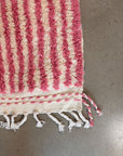Eine Nahaufnahme des LEY! Berberteppich - Pinq (1,20 x 2,00), rosa und weiß gestreift mit geflochtenen Quasten an einem Rand, ausgestellt auf einem glatten grauen Boden.