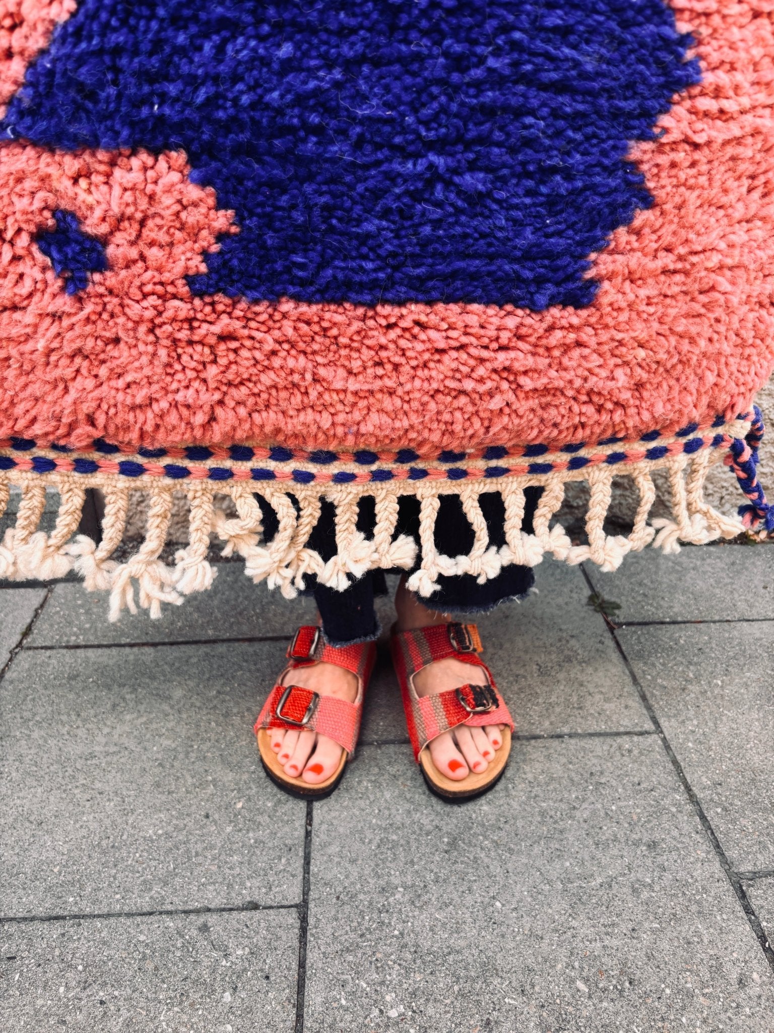 Eine Person in roten Sandalen steht neben dem Berber-Teppich „Farah“ (0,60 x 1,10 m) mit bunten Fransen von THE DANISH GIRL INTERIOR.