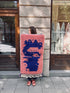 Eine Frau auf dem Bürgersteig hält den Berber-Teppich „THE DANISH GIRL INTERIOR“ – Farah, 0,60 x 1,10 m, in Rosa und Blau.