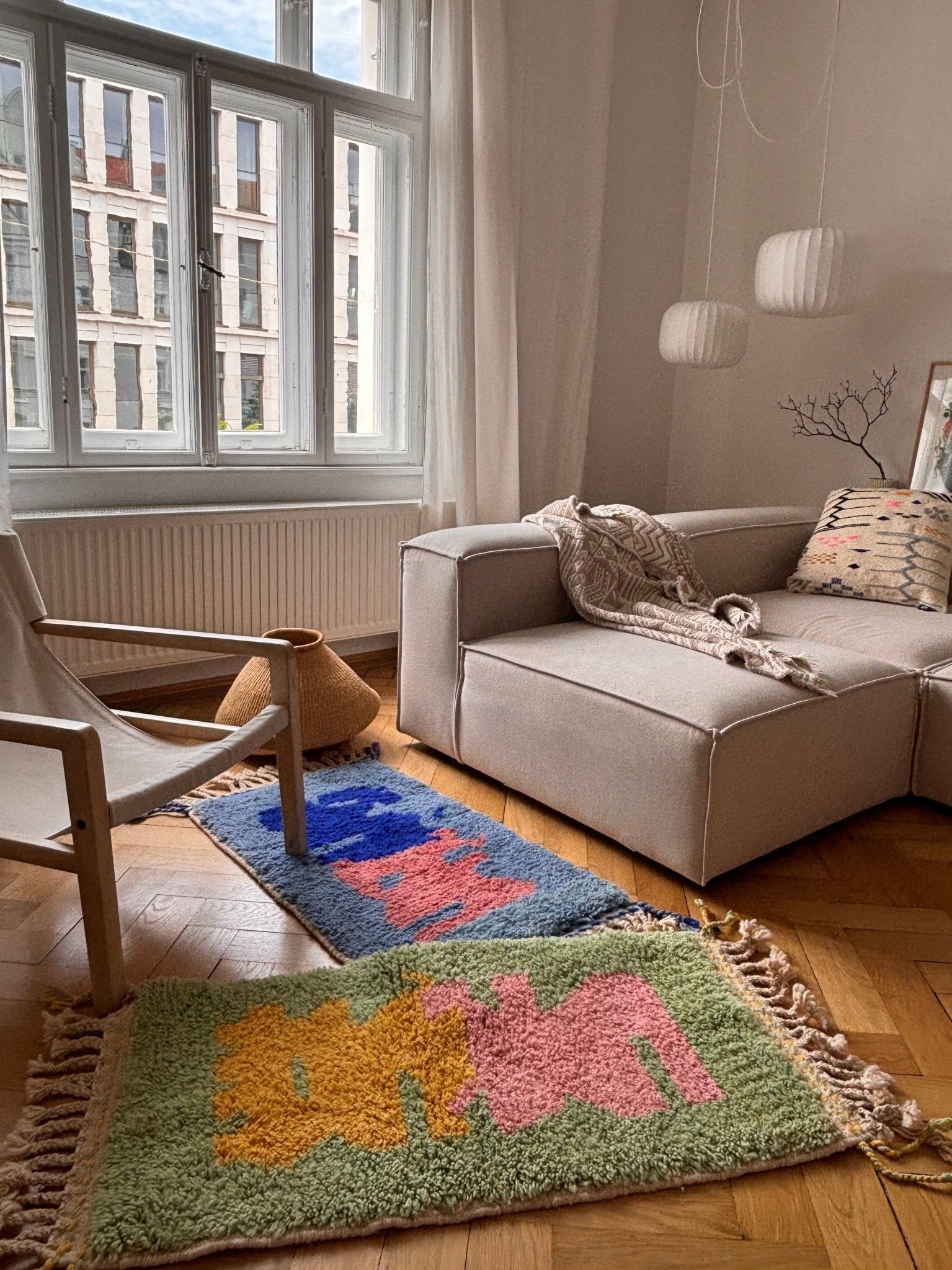 Das gemütliche Wohnzimmer mit einem beigen Sofa, einem modernen Sessel und zwei „Bariq“-Teppichen von THE DANISH GIRL INTERIOR sorgt für lebendige Farbakzente.