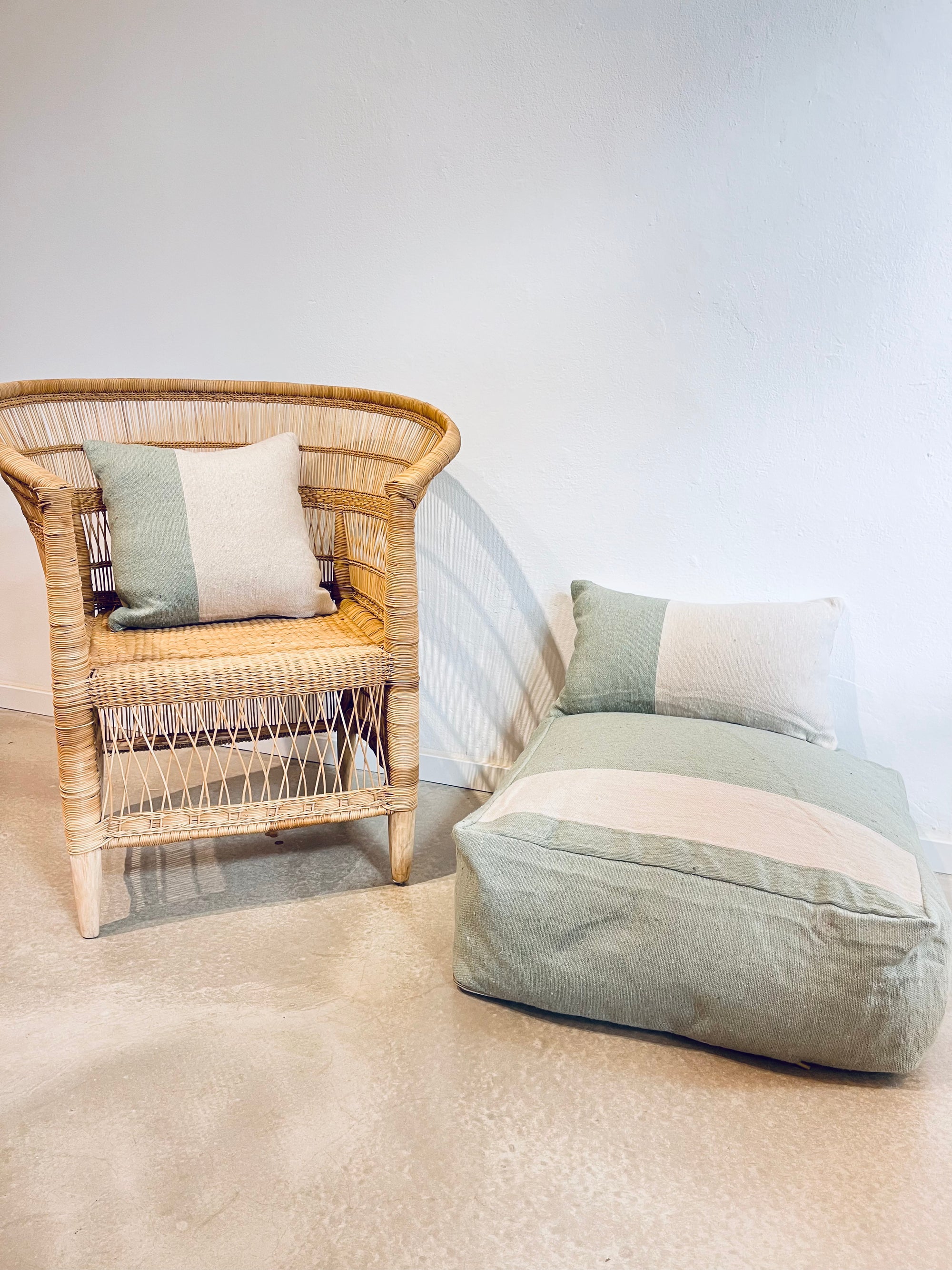 Ein Korbsessel mit gestreiftem Kissen sitzt neben dem LEY! Bodenkissen - Milena und einem Kissen, alles handgemacht aus weicher Baumwolle in neutralen und hellen Pastelltönen, vor einer schlichten weißen Wand auf einem hellen Boden.