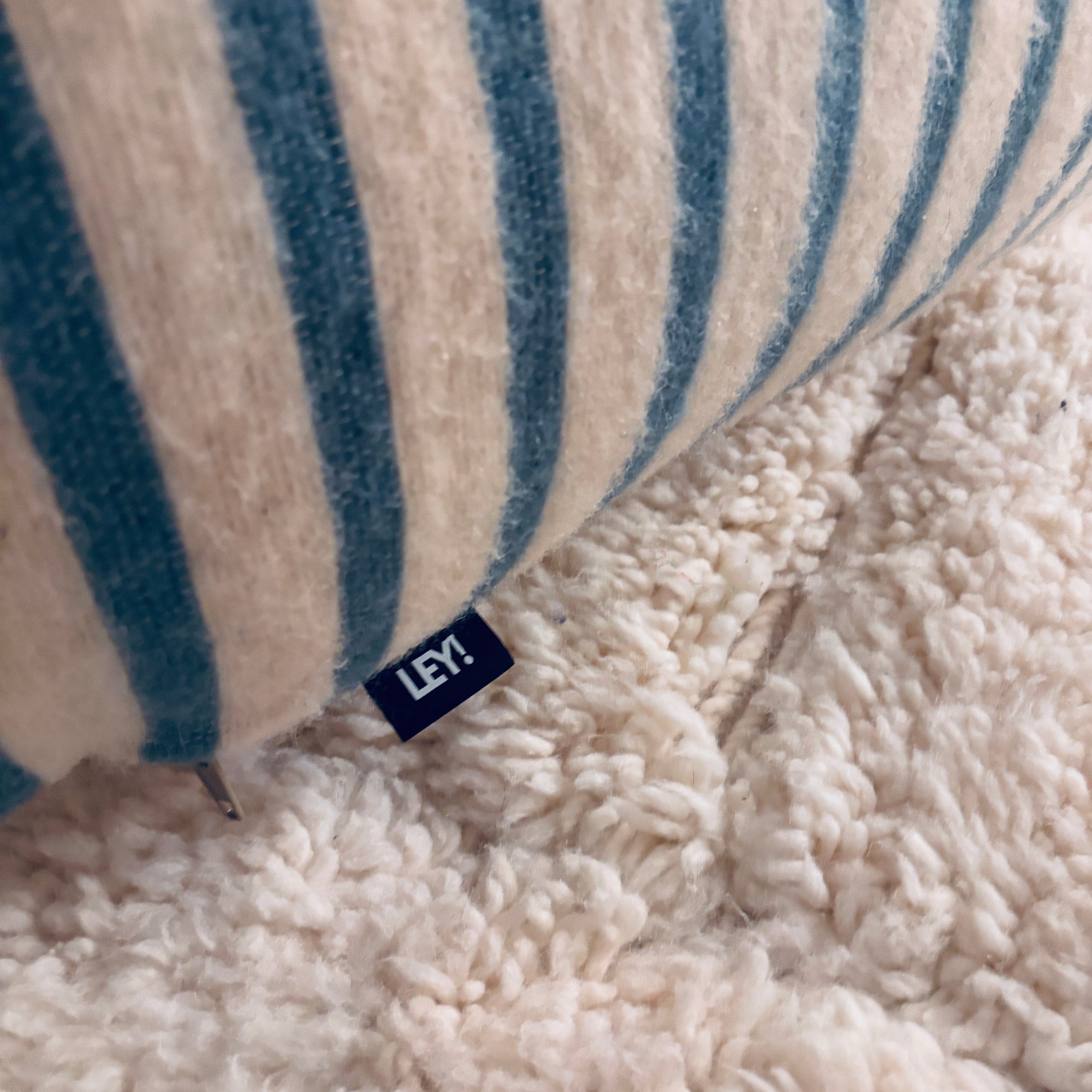 Eine Nahaufnahme des Bodenkissens - WINTER EDITION - Lagune von LEY! mit einem strukturierten cremefarbenen und blauen Streifendesign und einem kleinen "LEY!"-Anhänger, der auf einem weichen, cremefarbenen Zottelteppich liegt.