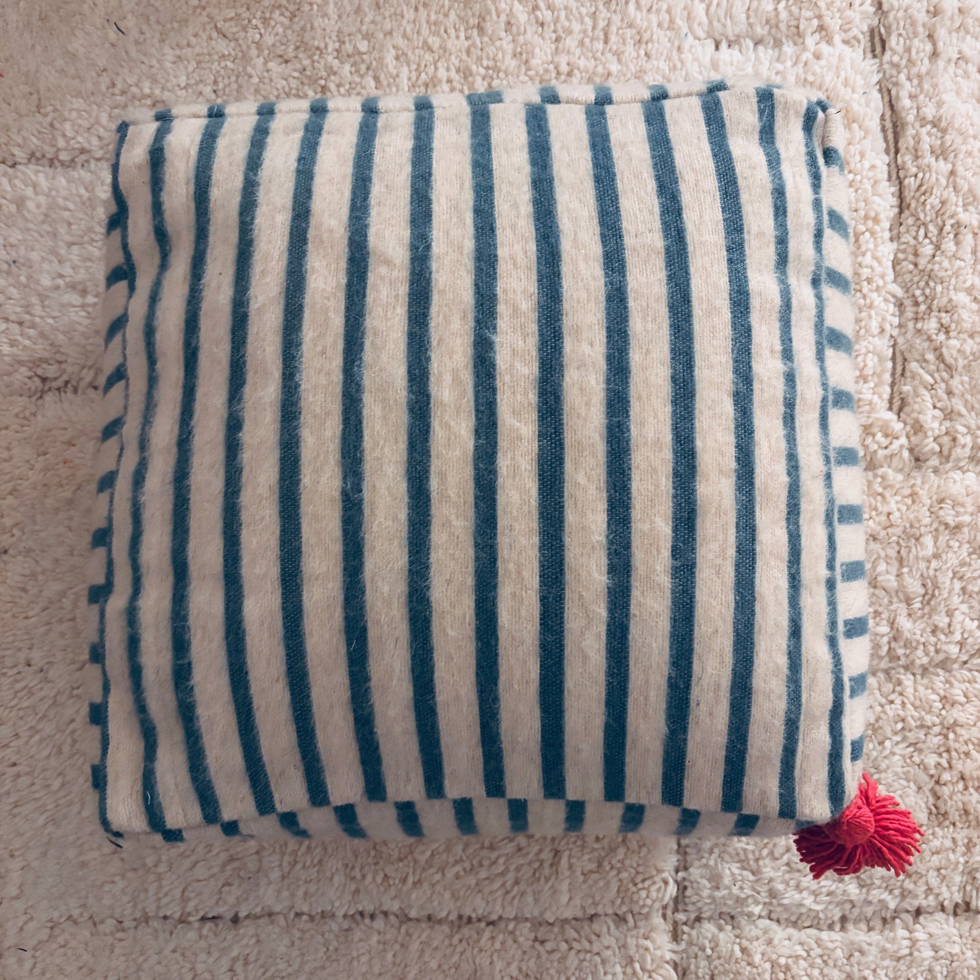 Ein quadratisches Bodenkissen - WINTER EDITION - Lagune von LEY! mit blauen und weißen Längsstreifen ruht auf einem beigen Teppich. Eine kleine rote Quaste akzentuiert eine Ecke - ideal für eine gemütliche saisonale Dekoration. Handgefertigt in Marrakesch.