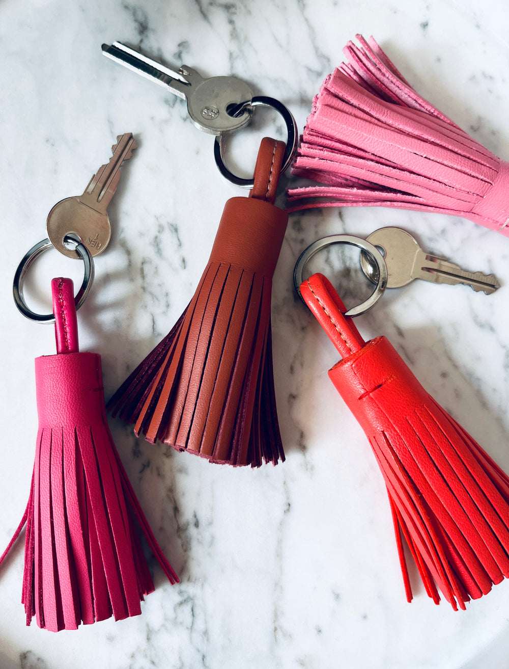 Drei Schlüssel mit Quastenanhängern aus rosa, braunem und rotem Leder liegen auf einer weißen Marmorfläche, zusammen mit einer zusätzlichen rosa Quaste ohne Schlüssel.