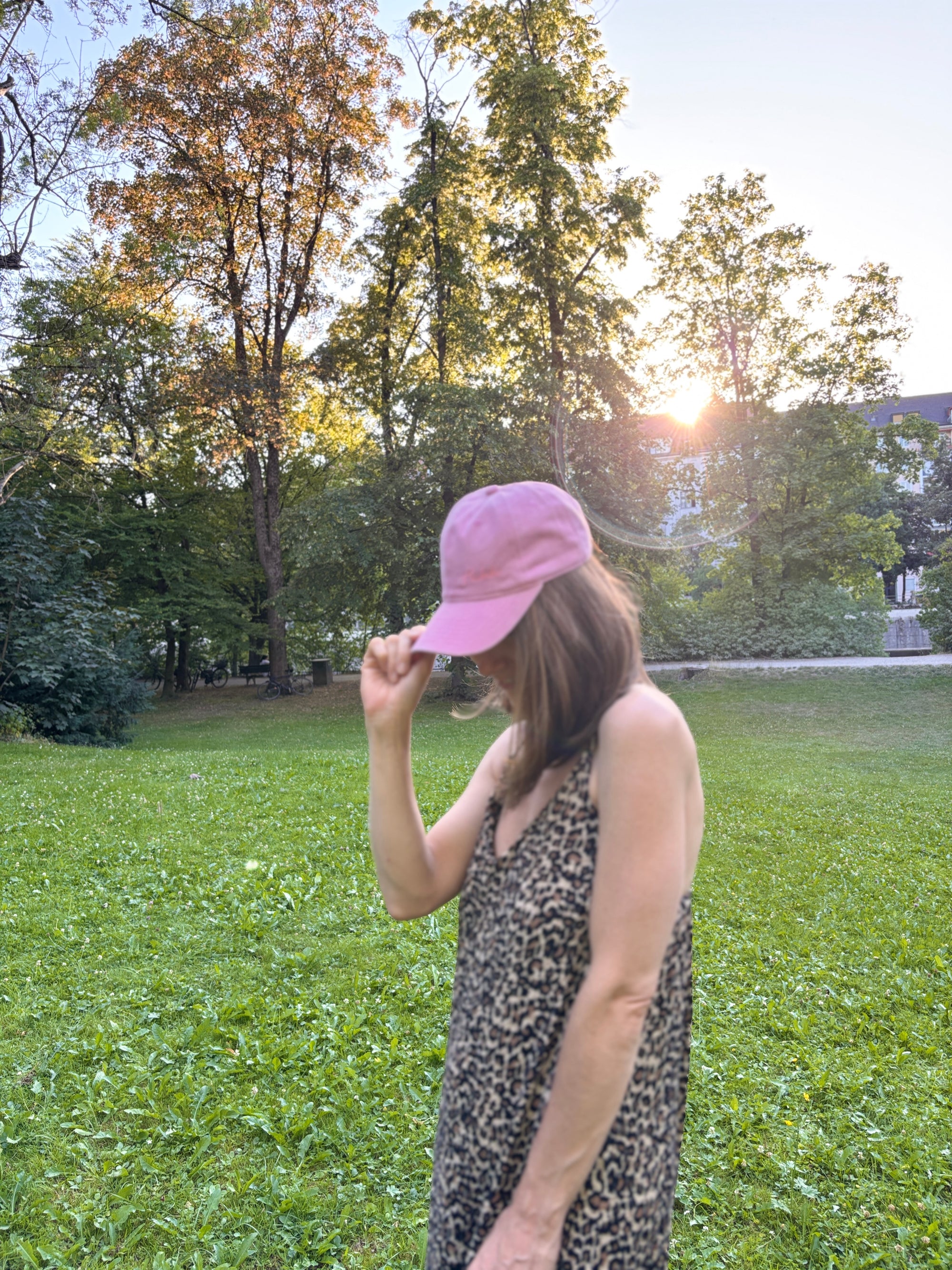 Eine Frau in einem Kleid mit Leopardenmuster und einer LEY! „Habibi“-Kappe in Blau steht auf einer Wiese, hinter den Bäumen scheint die Sonne.