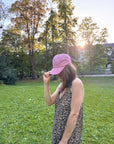 Eine Frau in einem Kleid mit Leopardenmuster und einer LEY! „Habibi“-Kappe in Blau steht auf einer Wiese, hinter den Bäumen scheint die Sonne.
