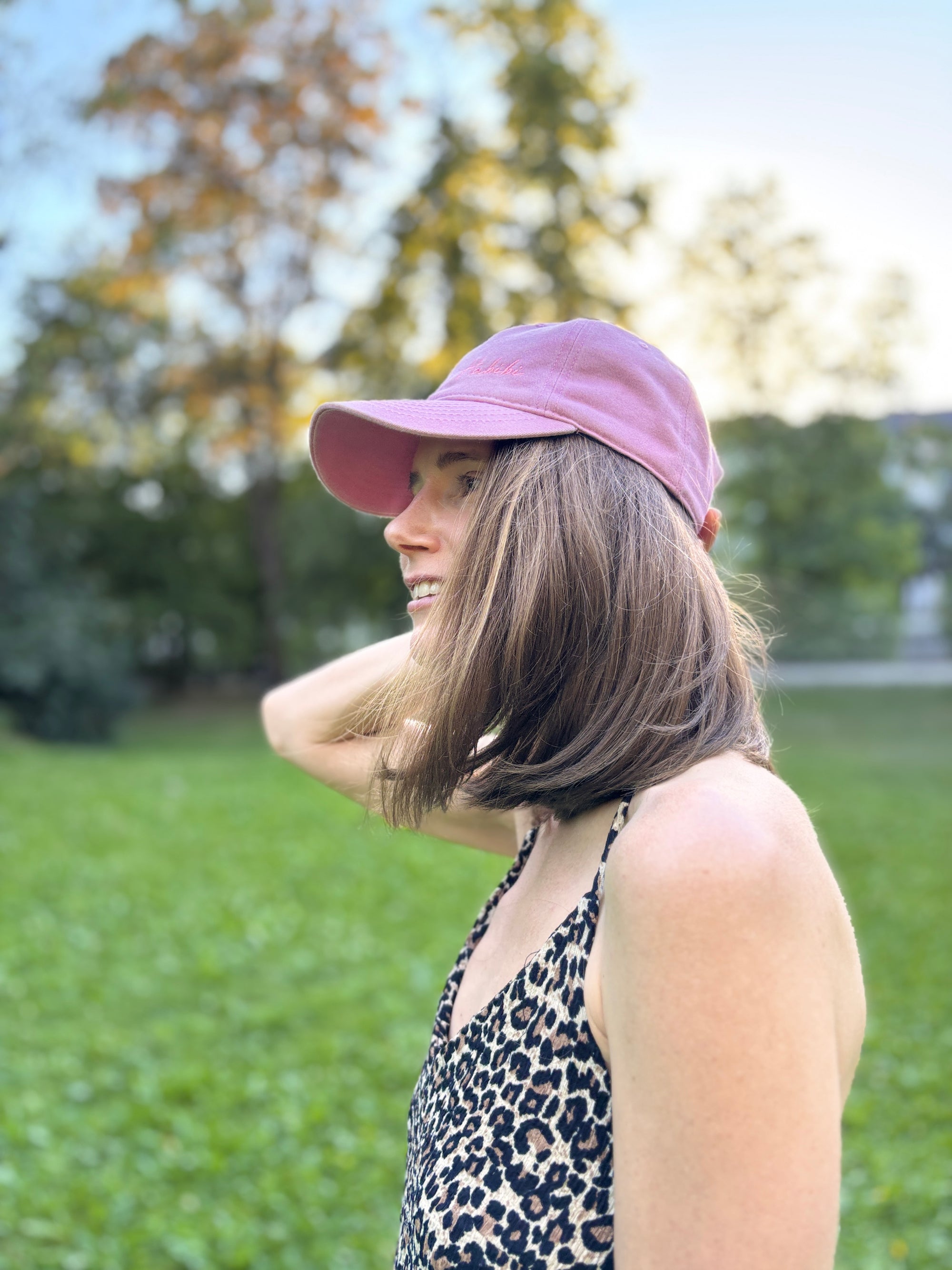 Eine Frau mit einer blauen LEY! „Habibi“-Kappe mit verstellbarem Verschluss lächelt auf dem Rasen, hinter ihr stehen Bäume.