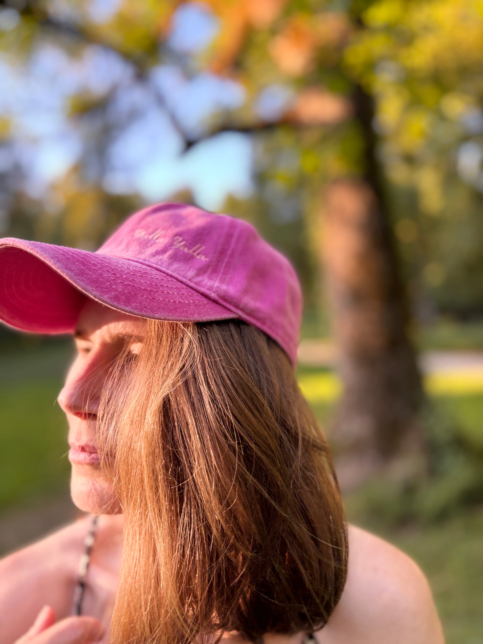 Eine Person mit langen braunen Haaren, die eine LEY! „Yalla Yalla“-Kappe in Lila trägt und im Freien vor einem verschwommenen grün-gelben Hintergrund steht.