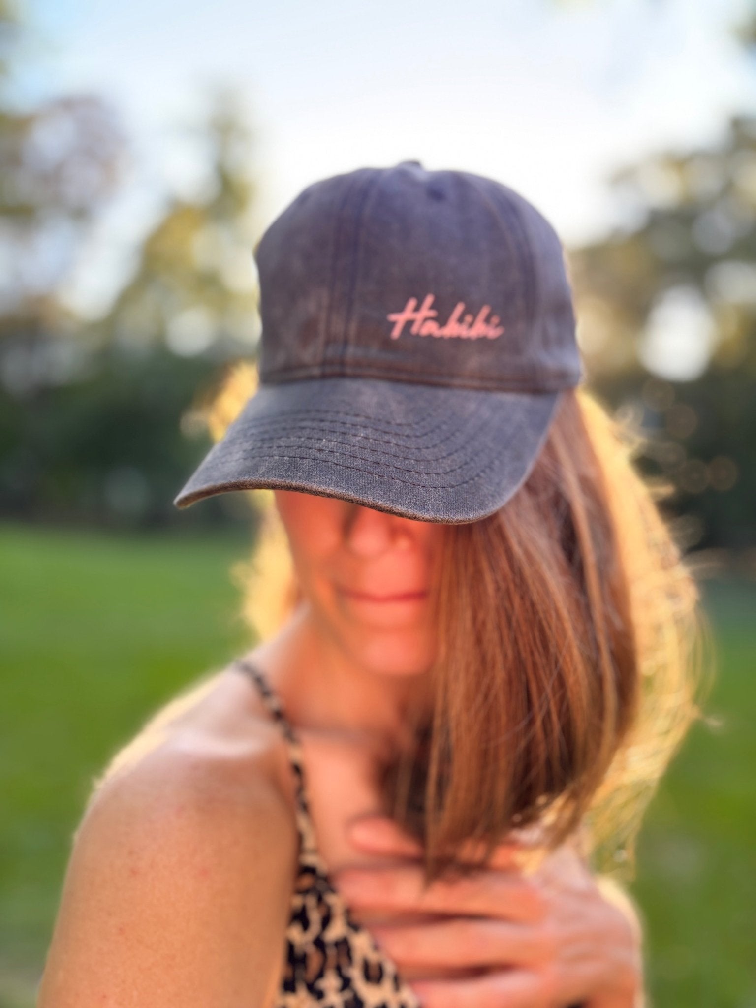 Eine Frau im Freien mit einer rosa LEY! „Habibi“-Kappe mit verstellbarem Riemen, im Hintergrund Sonnenlicht und Bäume.