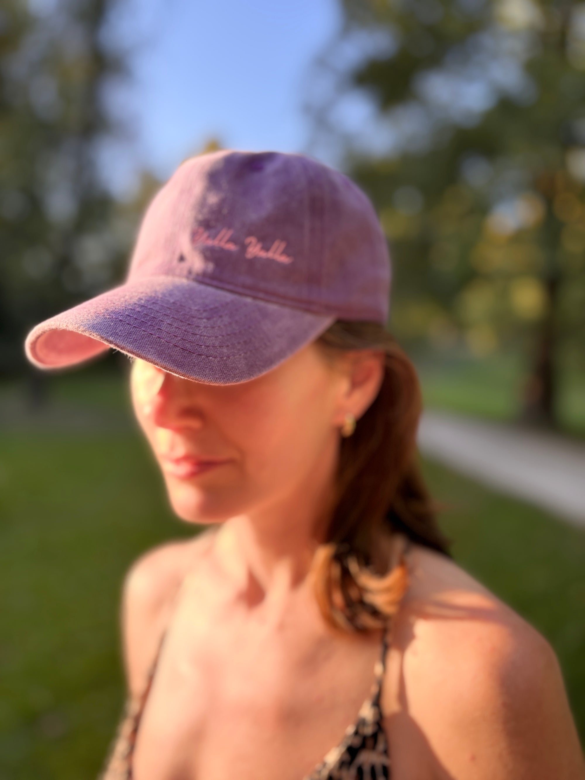 Eine Frau im Freien im Sonnenlicht, die die LEY! „Yalla Yalla“-Kappe in Grün trägt; hinter ihr sind Bäume und Gras sanft verschwommen.