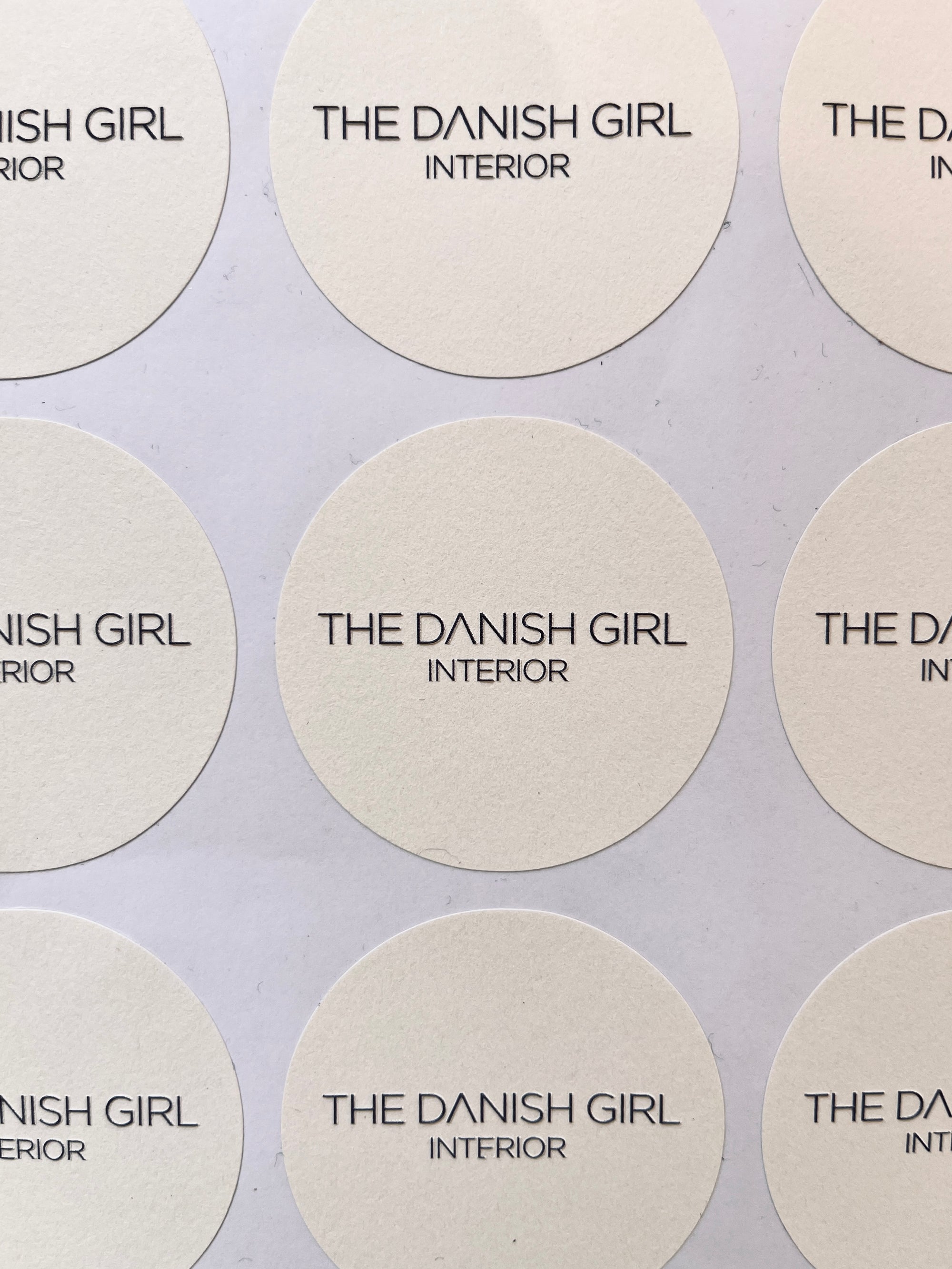 Ein runder, weißer LEY! STICKER mit "THE DANISH GIRL INTERIOR" in schwarz, angeordnet in einem Raster. Ideal als personalisierbarer Aufkleber oder Aufkleber mit Logo.