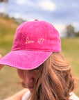 Eine Frau mit gewelltem Haar trägt draußen die „Love it“-Kappe in Rosa von LEY! und kreiert damit einen stilvollen Unisex-Look.
