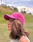 Eine Frau mit welligem braunem Haar trägt die LEY! „Love it“-Kappe in Rosa und ein weißes Oberteil, draußen vor dem Hintergrund von Gras und Bäumen.