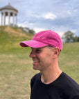 Ein Mann mit einer LEY!-Kappe mit der Aufschrift „Love it“ und einem rosa-schwarzen T-Shirt lächelt im Freien vor einem grasbewachsenen Hügel und einem Pavillon.