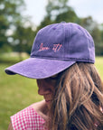 Frau mit einer „Love it“-Kappe von LEY! in Lila, draußen auf einer Wiese in einem rosa karierten Oberteil.