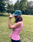 Eine Frau in einem rosa karierten Oberteil und einer „LEY! Love it“-Kappe in Grün steht in einem Park mit Rasenfläche und hält sich die Krempe ihrer Kappe fest.