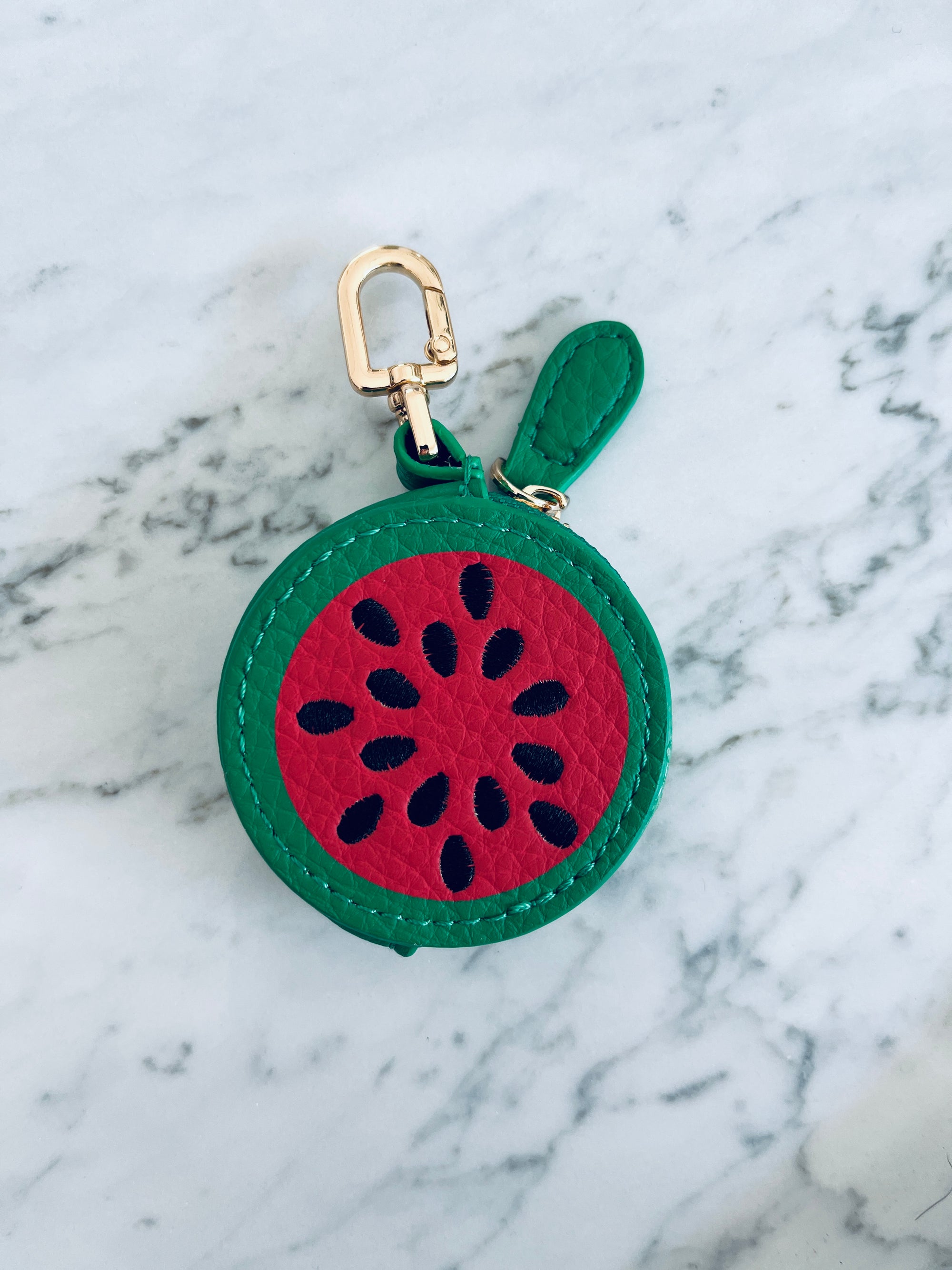 Der LEY! Schlüsselanhänger - Melone, ein kleines rundes Mini-Etui aus Kunstleder mit Reißverschluss in Wassermelonen-Design und goldener Befestigung, liegt auf einer hellen Marmoroberfläche.