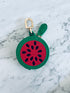 Der LEY! Schlüsselanhänger - Melone, ein kleines rundes Mini-Etui aus Kunstleder mit Reißverschluss in Wassermelonen-Design und goldener Befestigung, liegt auf einer hellen Marmoroberfläche.