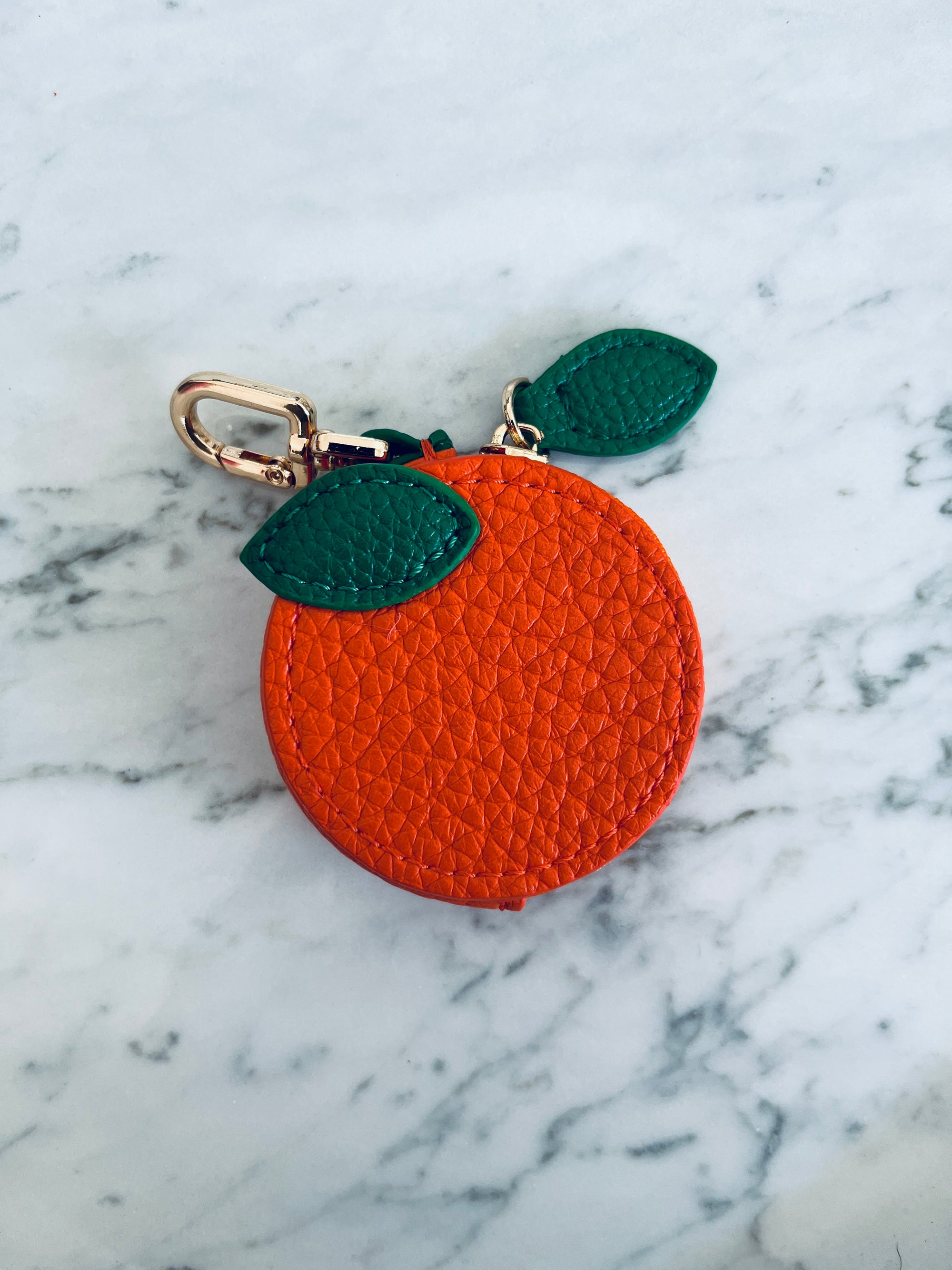 Runder orangefarbener Schlüsselanhänger - Etui von LEY! mit grünem Blattdetail und Schließe, auf einer weißen Marmoroberfläche.