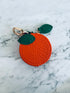 Runder orangefarbener Schlüsselanhänger - Etui von LEY! mit grünem Blattdetail und Schließe, auf einer weißen Marmoroberfläche.