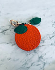 Runder orangefarbener Schlüsselanhänger - Etui von LEY! mit grünem Blattdetail und Schließe, auf einer weißen Marmoroberfläche.