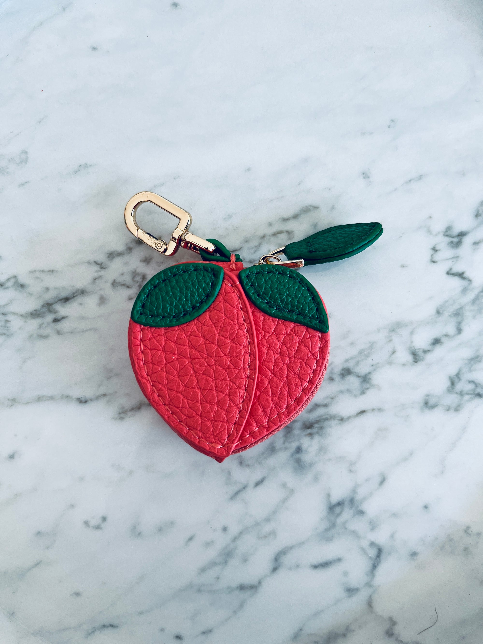 Der LEY! Schlüsselanhänger - Pfirsich ist ein kleines, pfirsichförmiges Portemonnaie aus strukturiertem rosa Mini Etui Kunstleder mit grünen Blattakzenten und einer goldenen Schließe, das auf einer weißen Marmoroberfläche präsentiert wird.