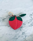 Der LEY! Schlüsselanhänger - Pfirsich ist ein kleines, pfirsichförmiges Portemonnaie aus strukturiertem rosa Mini Etui Kunstleder mit grünen Blattakzenten und einer goldenen Schließe, das auf einer weißen Marmoroberfläche präsentiert wird.