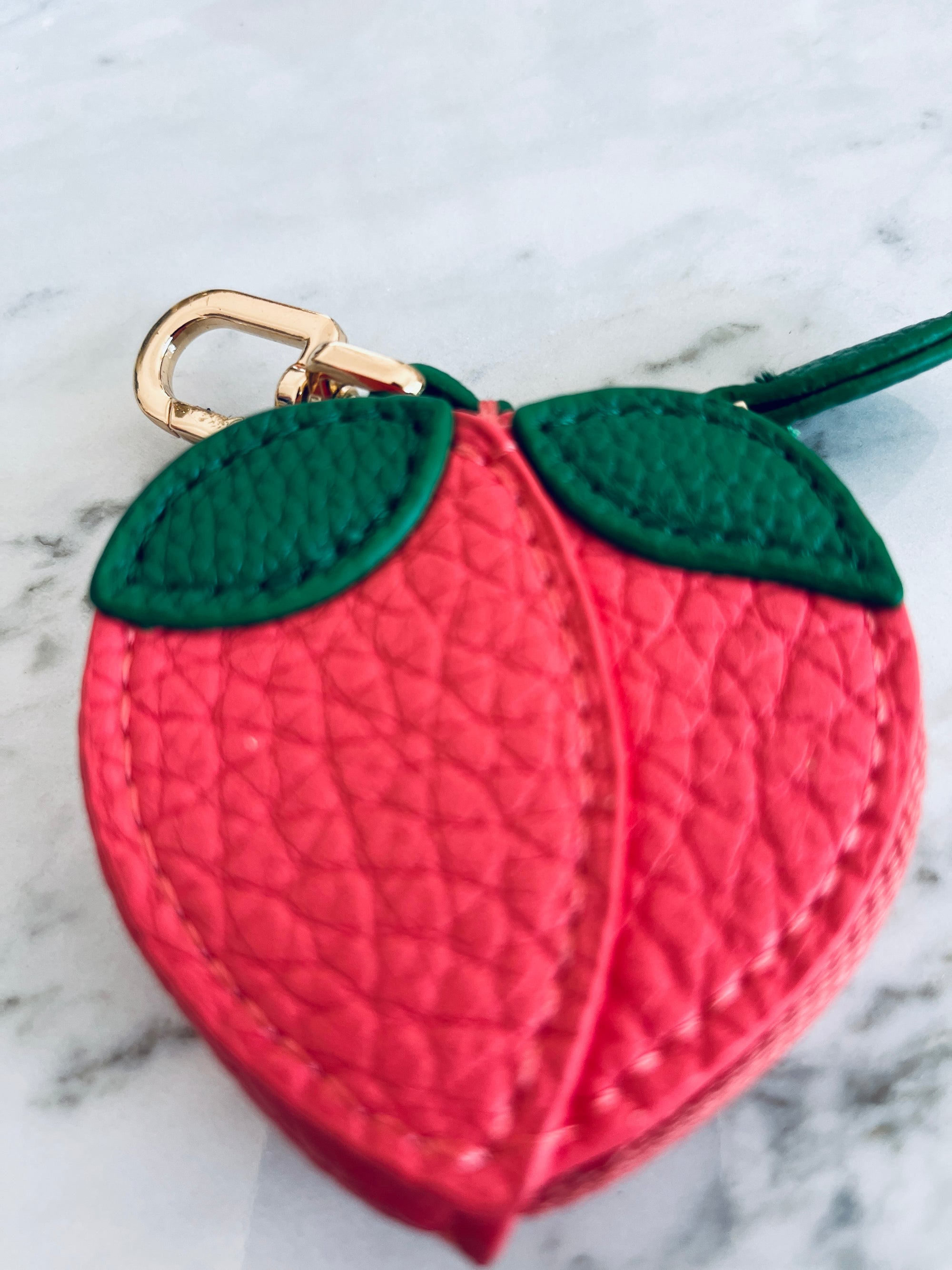 Der Schlüsselanhänger - Pfirsich von LEY! ist ein kleiner, strukturierter, rosafarbener Anhänger in Pfirsichform mit grünen Blättern und einer goldenen Schließe - ideal als Mini-Etui aus Kunstleder für Ihre Essentials.