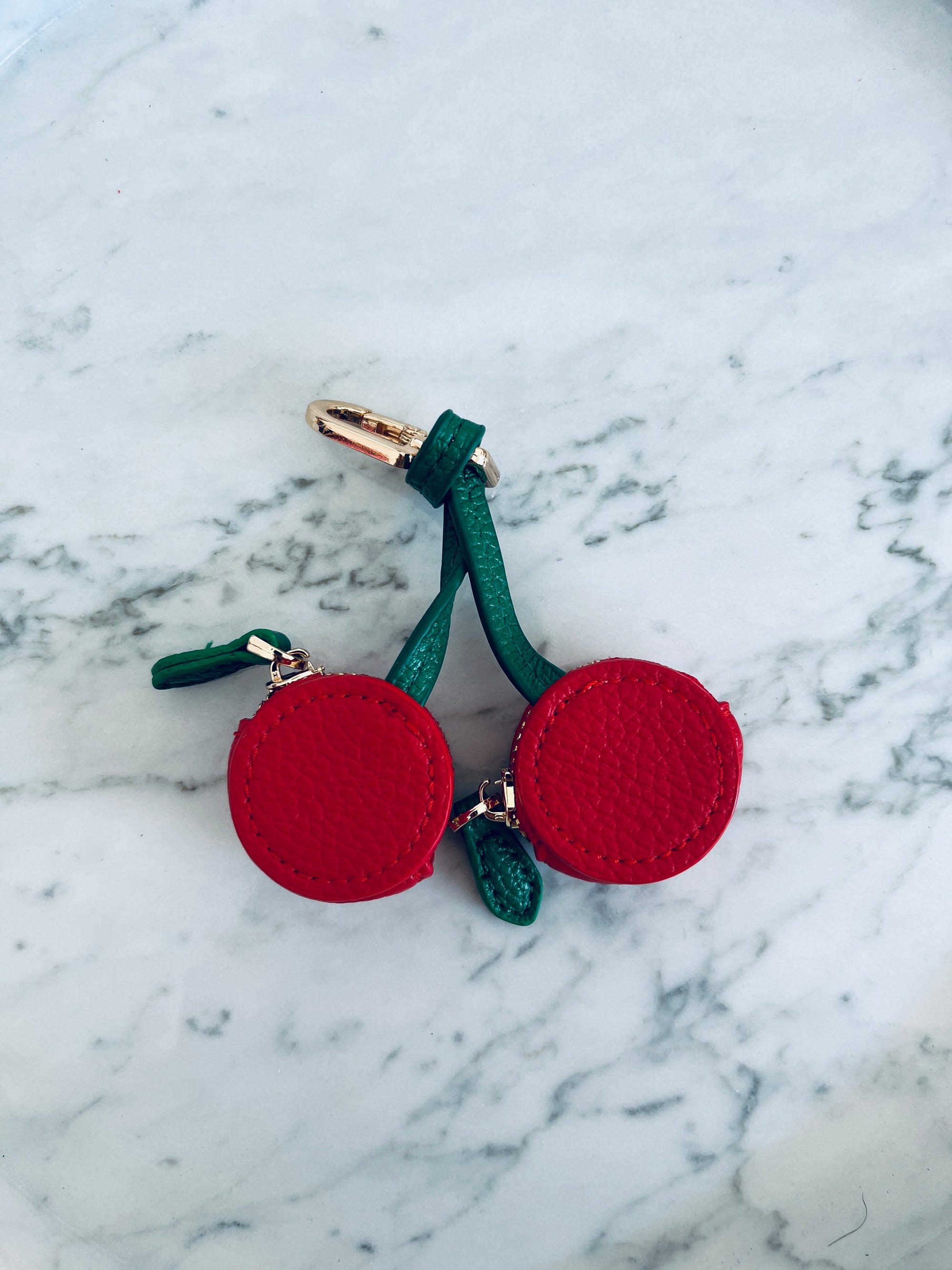 Der Schlüsselanhänger - Kirsche von LEY! zeigt zwei rote, runde Charms aus Mini Etui Kunstleder an grünen Stielen mit goldener Schlaufe, stilvoll auf Marmor präsentiert.