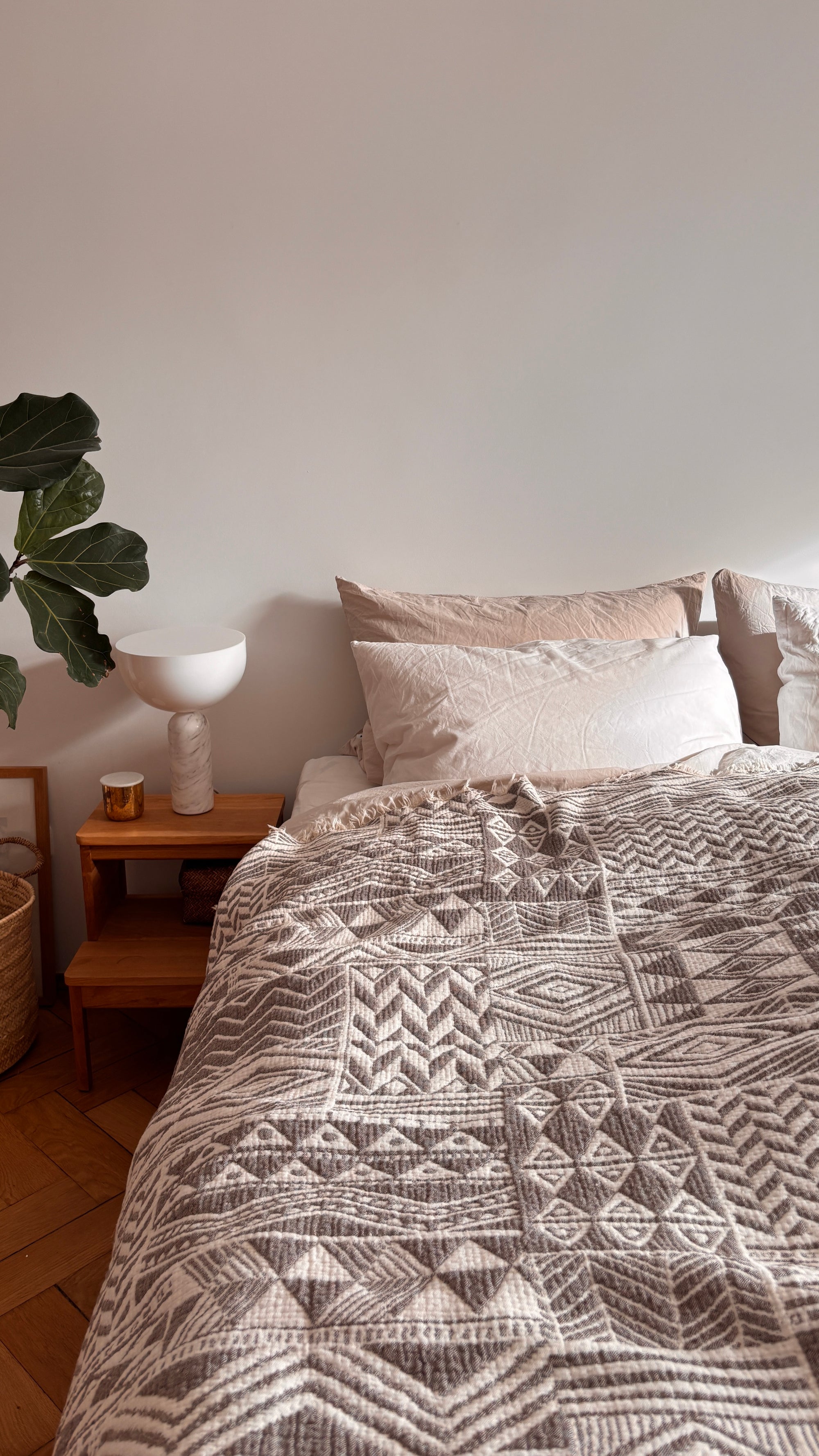 Ein gemütliches Schlafzimmer mit einem ordentlich gemachten Bett mit gemusterter Bettwäsche und der LEY! CALMA Musselin Decke in Grau-Weiß (1,50 x 2,40 m), die für ihren atmungsaktiven Komfort bekannt ist, sowie neutralen Kissen, einem hölzernen Nachttisch mit Lampe, Kerze und Pflanze - alles vor einer weißen Wand.