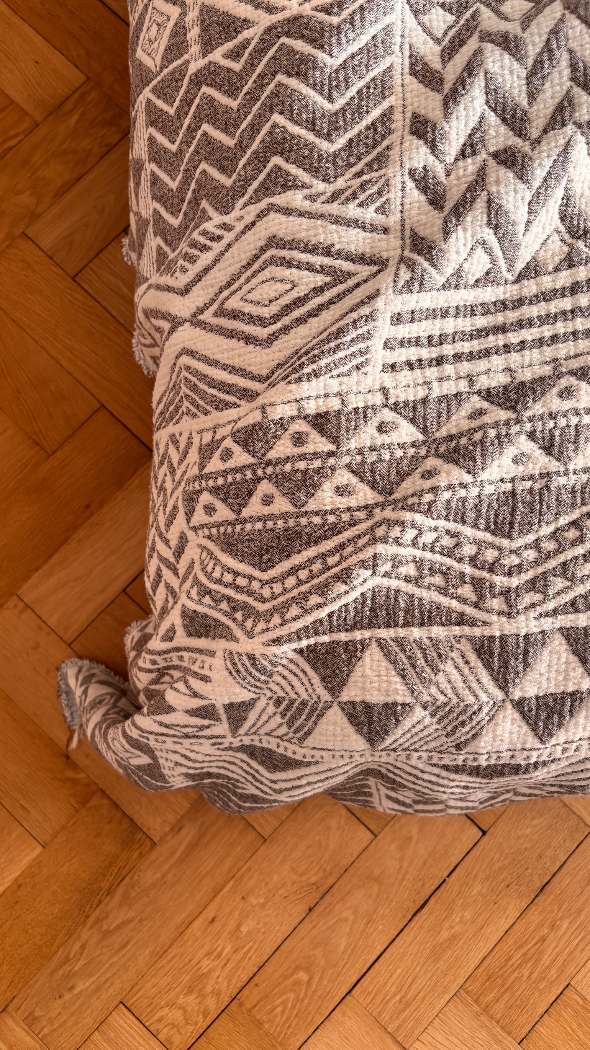 Eine Nahaufnahme der CALMA Musselin Decke in Grau-Weiß (1,50 x 2,40 m) von LEY! im stilvollen Boho-Design, drapiert auf einer Fläche mit Holzparkett im Hintergrund.