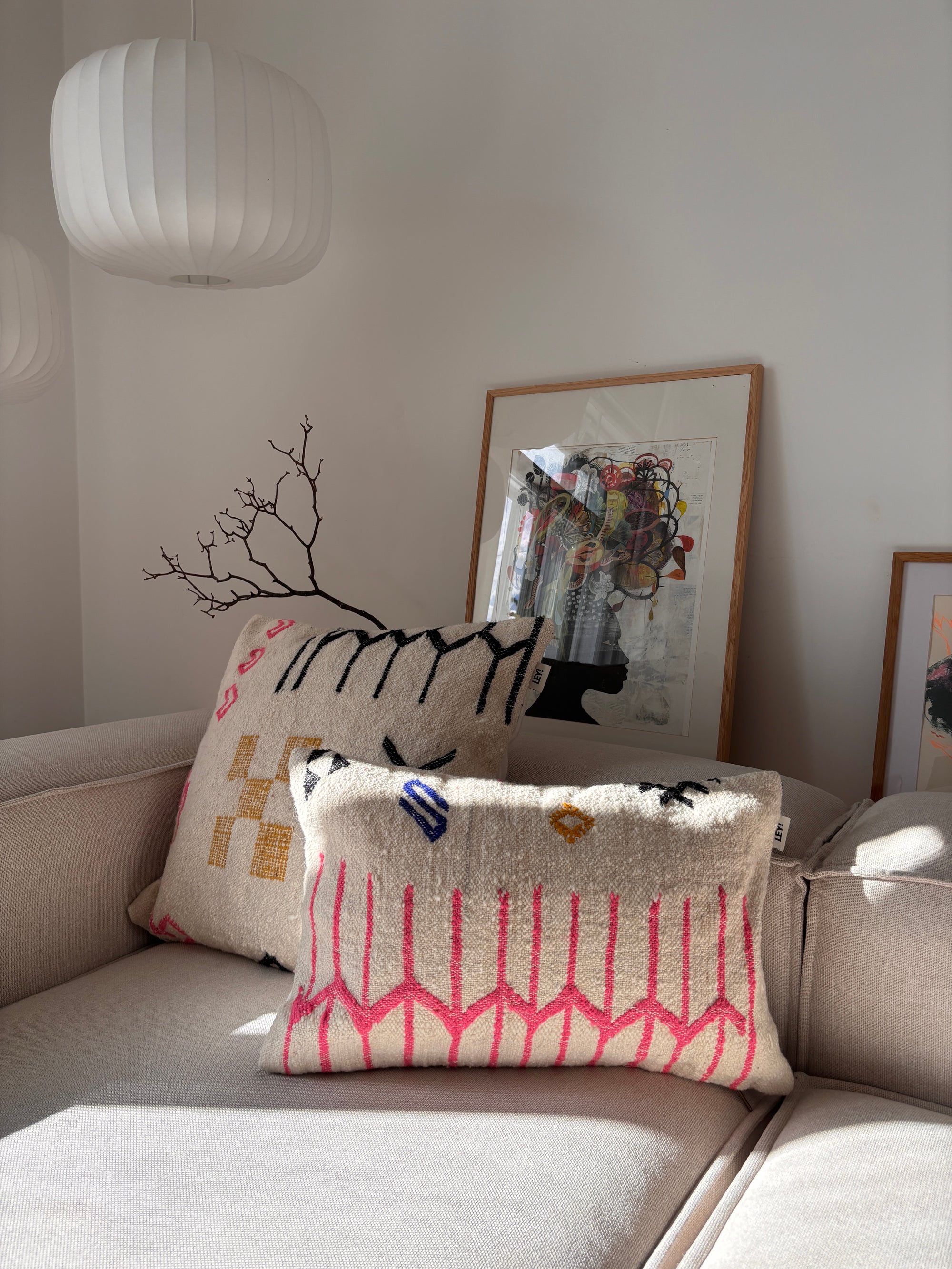 Zwei LEY!-Kissen – Colorful Line – 40 x 60 von LEY! liegen auf einem beigen Sofa, umgeben von Kunstwerken und einer Lampe, in einem sonnendurchfluteten Raum.