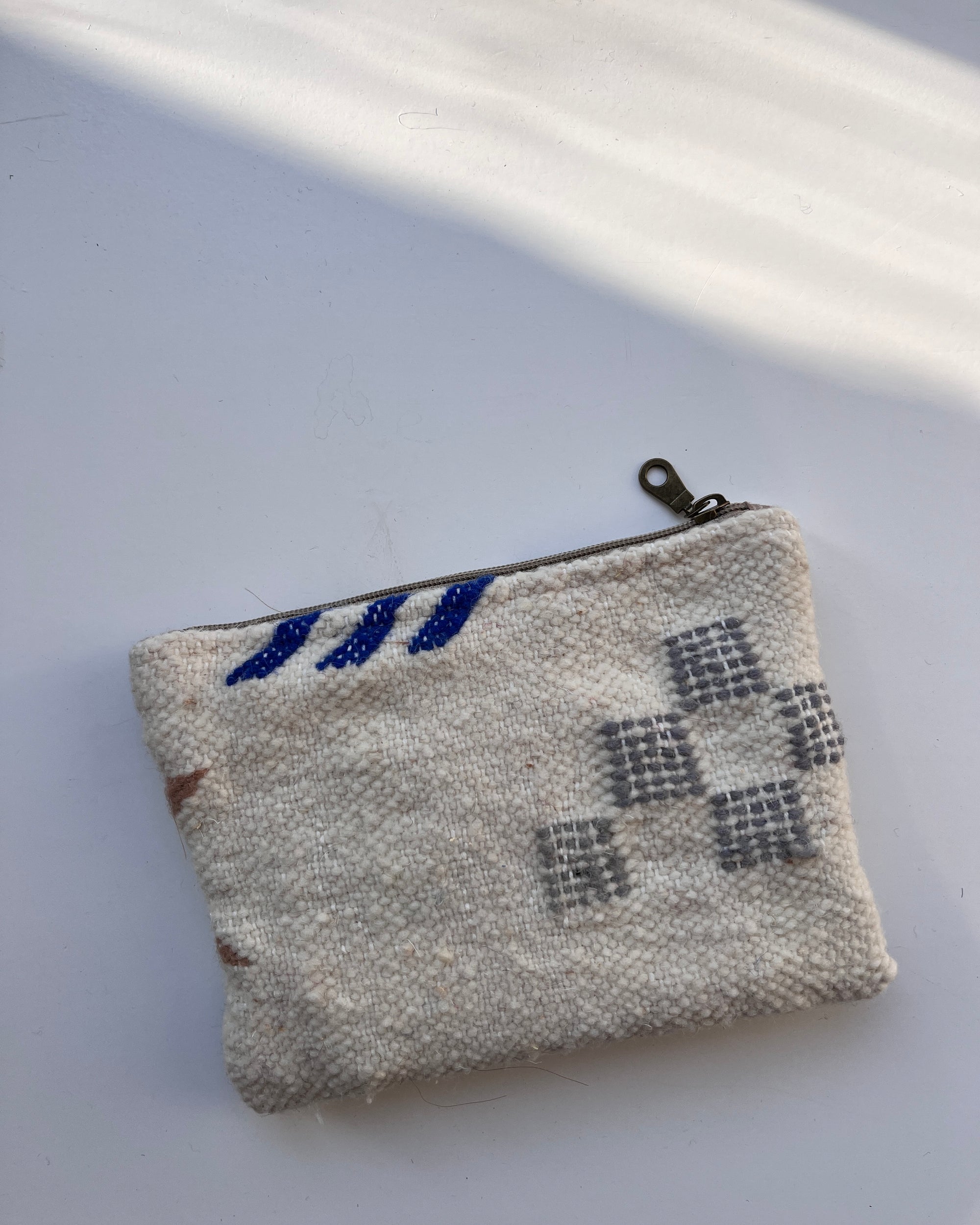 Eine kleine, strukturierte weiße Tasche von LEY! mit Reißverschluss und blaugrauen Stickmustern ruht auf einer weißen Fläche, die in diagonales Sonnenlicht getaucht ist.
