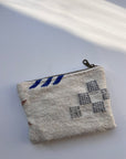 Eine kleine, strukturierte weiße Tasche von LEY! mit Reißverschluss und blaugrauen Stickmustern ruht auf einer weißen Fläche, die in diagonales Sonnenlicht getaucht ist.