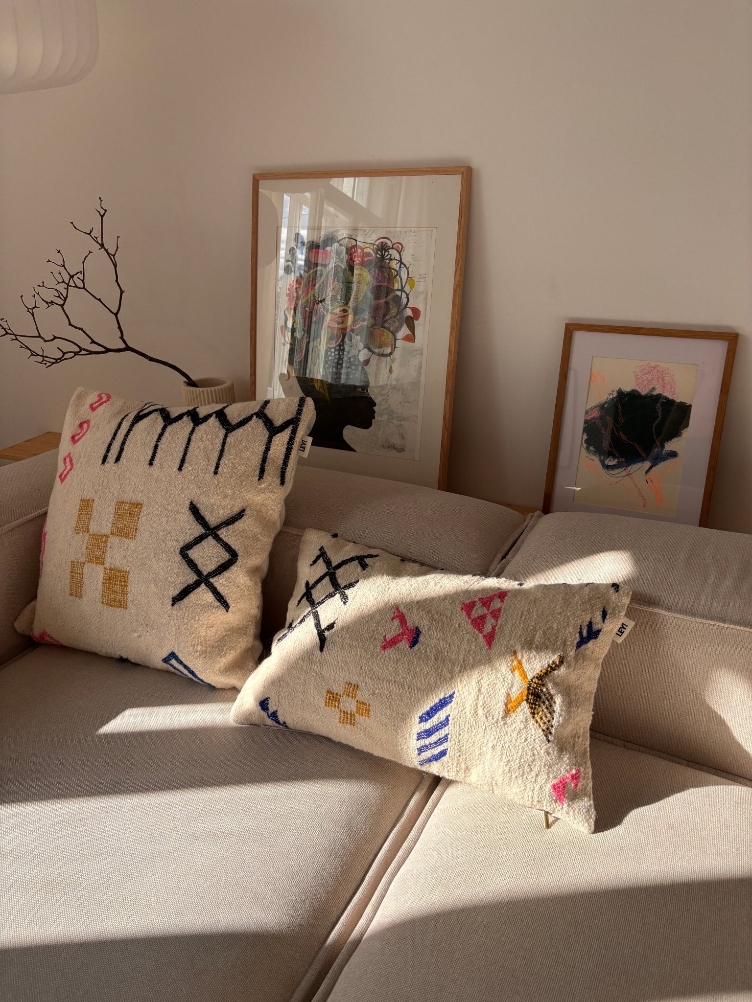 Zwei LEY!-Kissen – „Colorful Line“ 40 x 60 auf einem beigen Sofa, Sonnenstrahlen, die hereinscheinen, gerahmte Kunstwerke und ein Ast im Hintergrund.