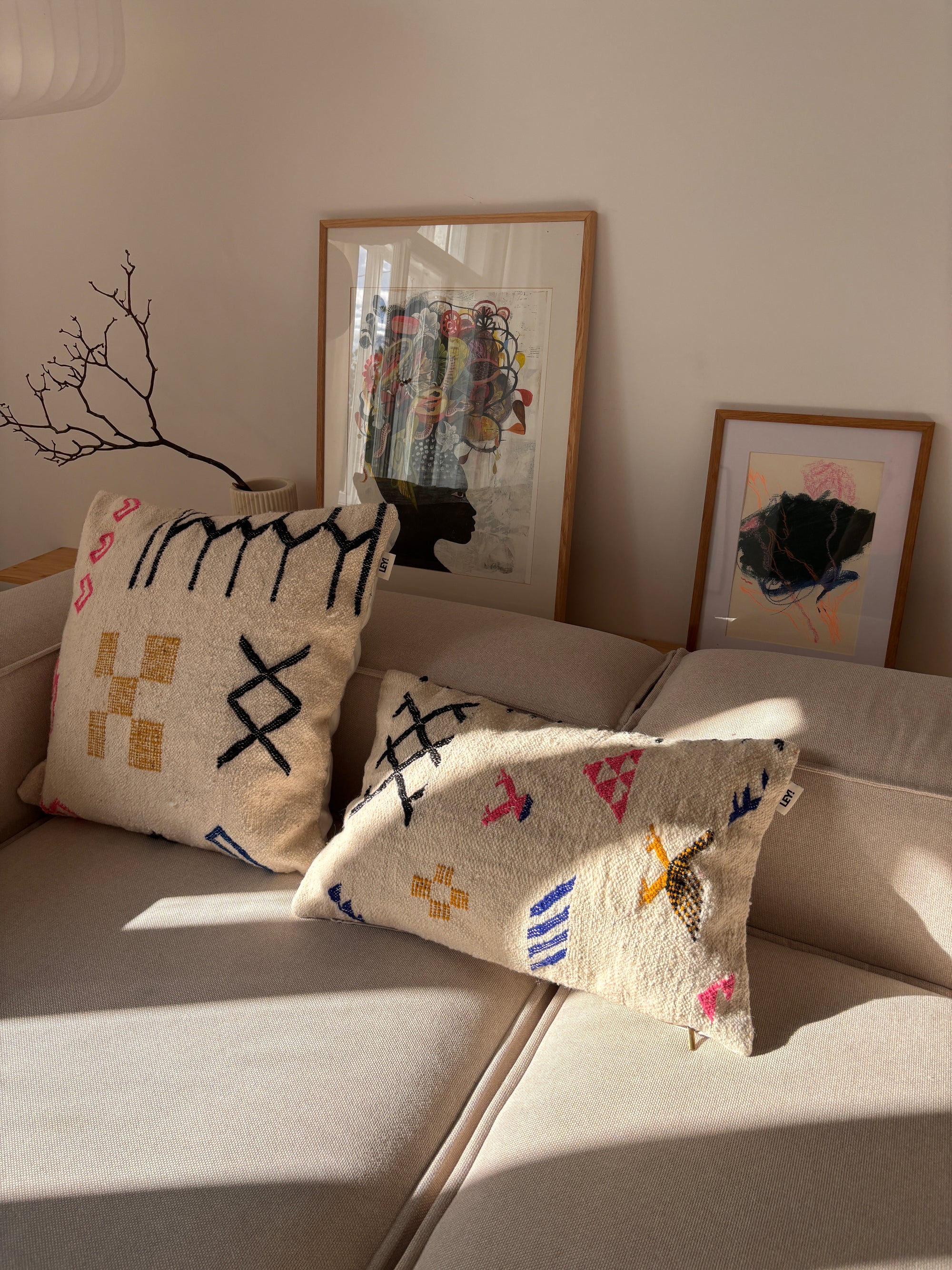 Zwei LEY!-Kissen – „Colorful Line“ 40 x 60 auf einem beigen Sofa, Sonnenstrahlen, die hereinscheinen, gerahmte Kunstwerke und ein Ast im Hintergrund.