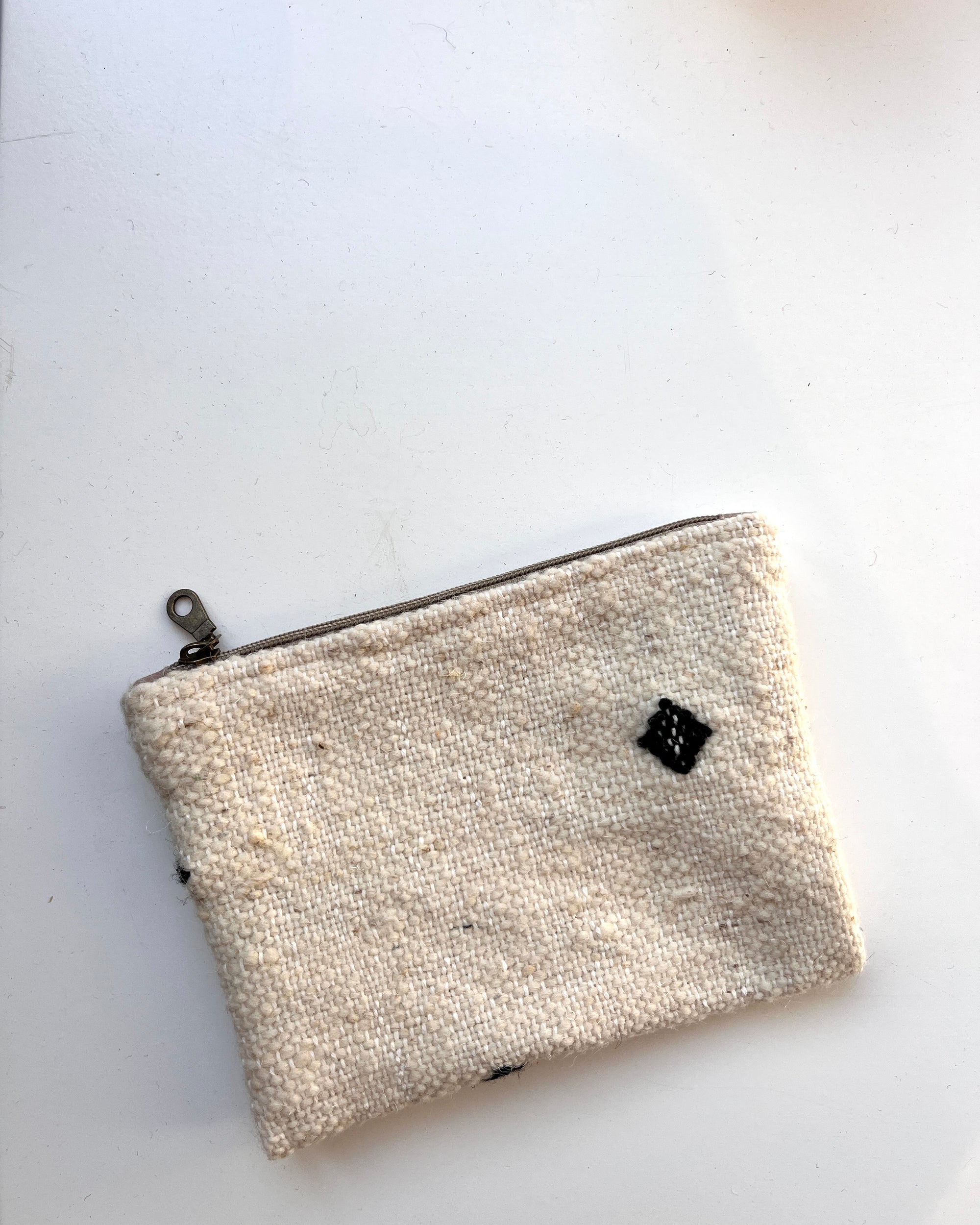 Die kleine, rechteckige Tasche von LEY! aus Schurwolle ist off-white, handgefertigt, mit Reißverschluss oben und schwarzen Quadrat-Akzenten und liegt auf einer weißen Oberfläche.