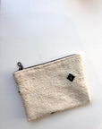 Die kleine, rechteckige Tasche von LEY! aus Schurwolle ist off-white, handgefertigt, mit Reißverschluss oben und schwarzen Quadrat-Akzenten und liegt auf einer weißen Oberfläche.