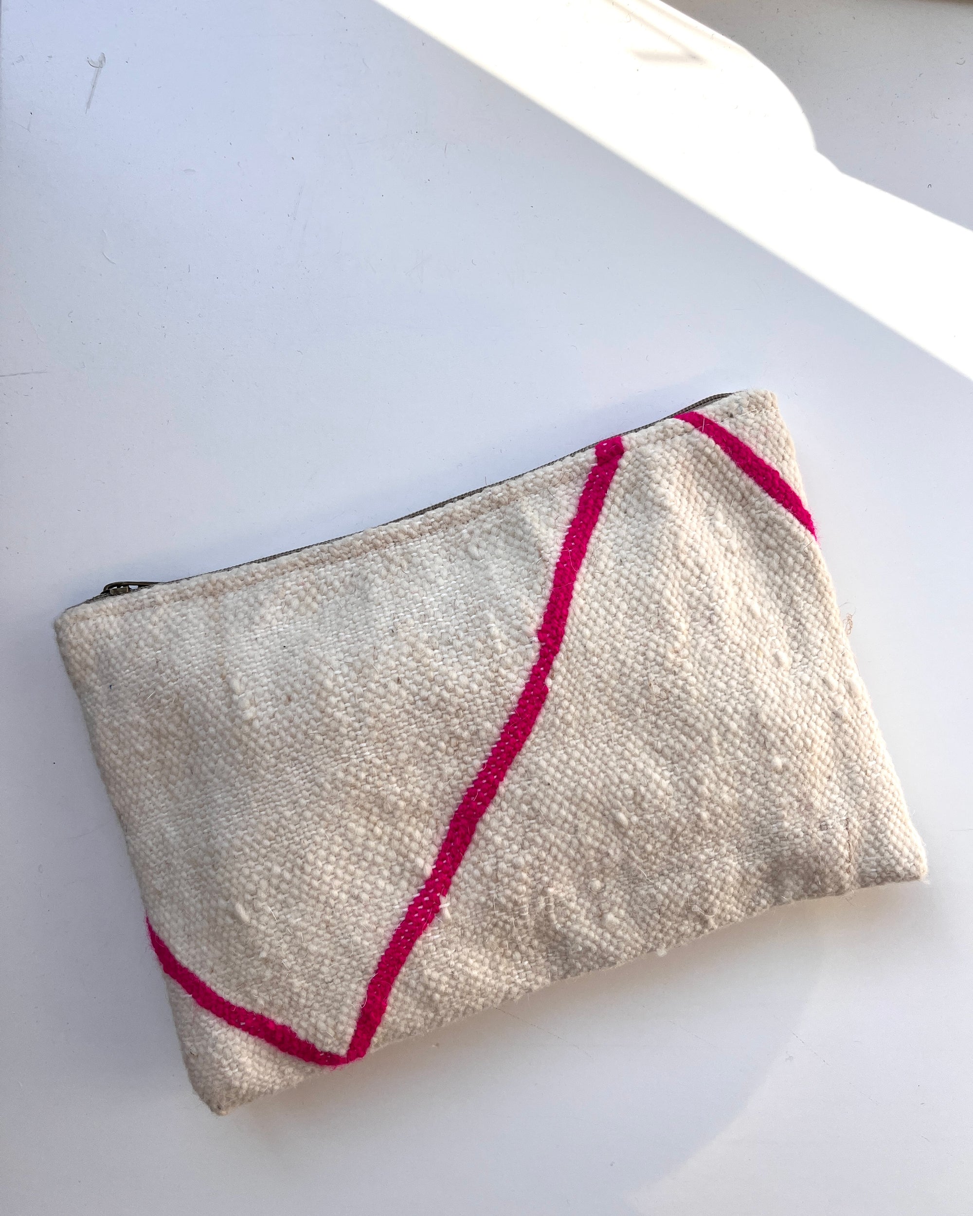 Die LEY! Tasche - Mina - 20X30 ist eine kleine, strukturierte, beigefarbene, handgefertigte Tasche mit Reißverschluss und unregelmäßigen, hellrosa Linien, die auf einer weißen Oberfläche mit sanften Sonnenlichtschatten abgebildet ist.