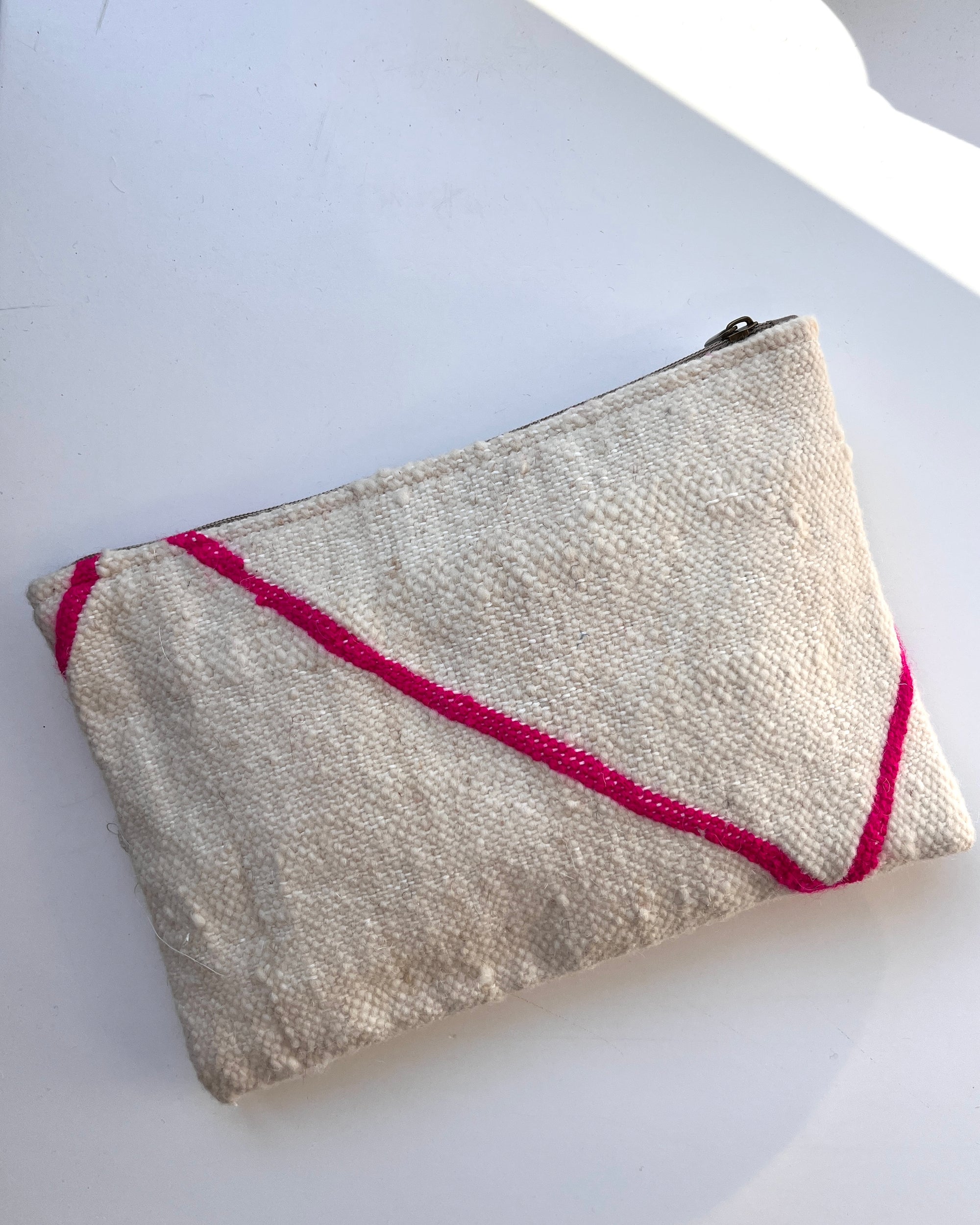 Die cremefarbene, strukturierte Tasche - Mina - 20X30 von LEY! aus Schurwolle hat leuchtend rosa Diagonalstreifen und ist in liebevoller Handarbeit gefertigt. Abgebildet ist sie auf einer weißen Fläche mit einem Schatten in der rechten oberen Ecke.