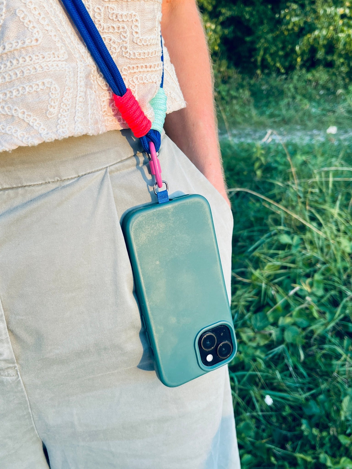 Jemand mit einem grünen Handy im LEY! Lange Handykette „LINA“ – Beere steht im Gras, in einer hellen Hose und einem weißen Oberteil.